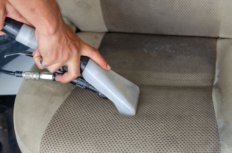 Close-up of a professional cleaner's gloved hand operating a steam cleaning machine on a dark carpeted surface, likely within a residential or commercial setting. The machine’s nozzle releases a jet
